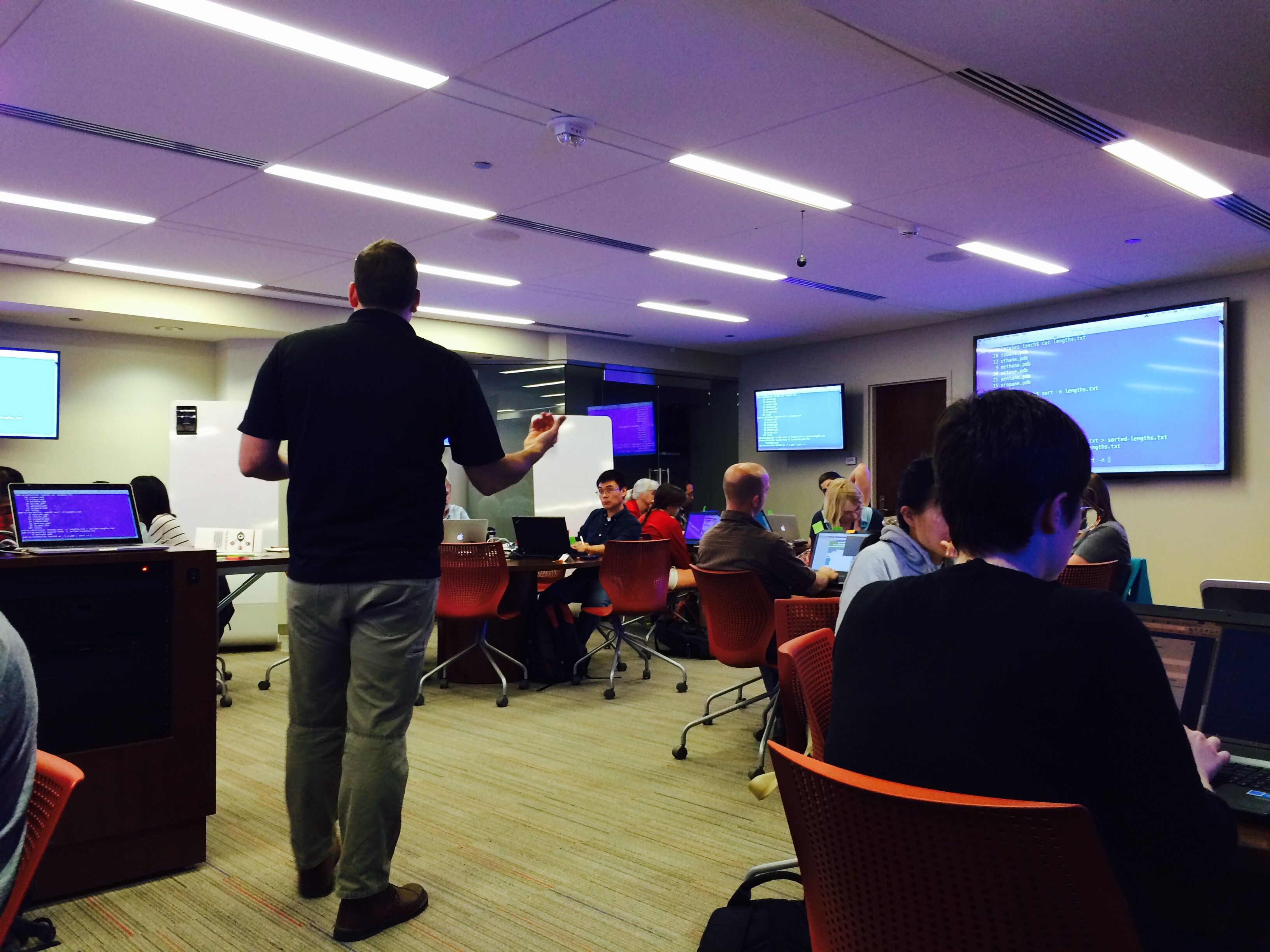 An instructor teaching a library workshop in an active learning classroom with multiple screens on the walls and people at tables surrounding the instructor