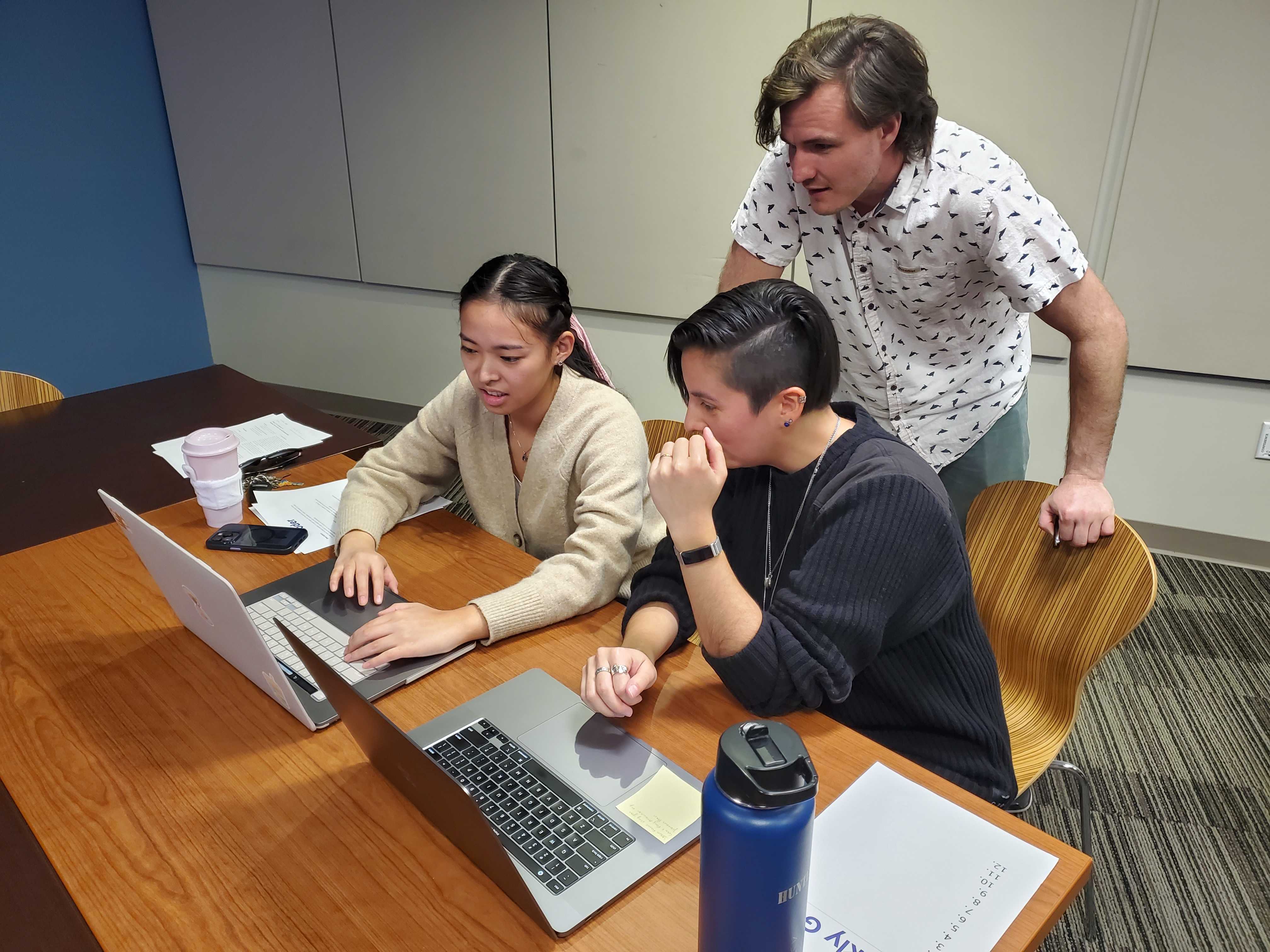 A grouping of three people. Two are sitting and one is standing. All three are looking at the laptops in front of them.
