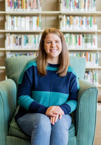 Hallie Macom smiling and sitting in a green chair with books on a shelf behind her.