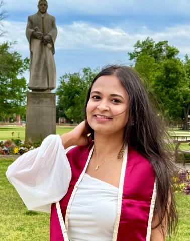 A woman in graduation attire smiles in a park with a statue in the background. She wears a white top and maroon sash.